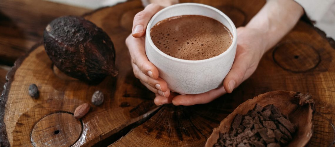 natuurlijke elementen van een traditionele “cacao ceremonie” oproept, waarbij de nadruk ligt op het verbinden met de spirituele en helende eigenschappen van pure, ceremoniële cacao. De afbeelding toont een paar handen die een witte keramische kop vasthouden, gevuld met een rijk, schuimend cacaodrankje, rustend op een houten oppervlak. In de buurt liggen een cacaovrucht en verschillende cacaonibs, wat de aardse en natuurlijke elementen van een traditionele "cacao ceremonie" oproept, waarbij de nadruk ligt op het verbinden met de spirituele en helende eigenschappen van pure, ceremoniële cacao.