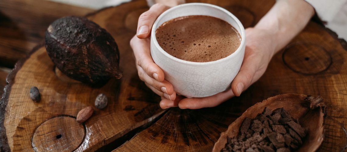 Hot handmade ceremonial cacao in white cup. Woman hands holding craft cocoa, top view on wooden table. Organic healthy chocolate drink prepared from beans, no sugar. Giving cup on ceremony, cozy cafe